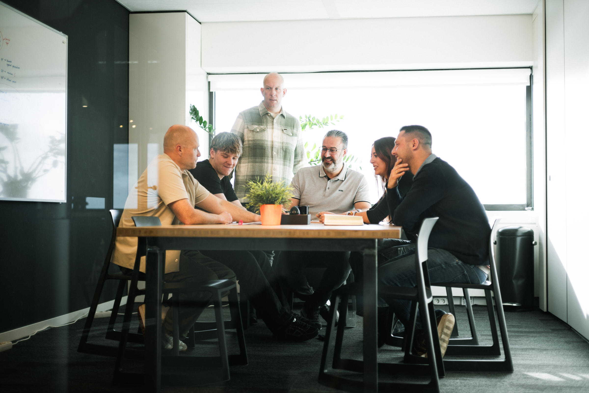 Six people having a meeting around a table in a bright office with a whiteboard and window.