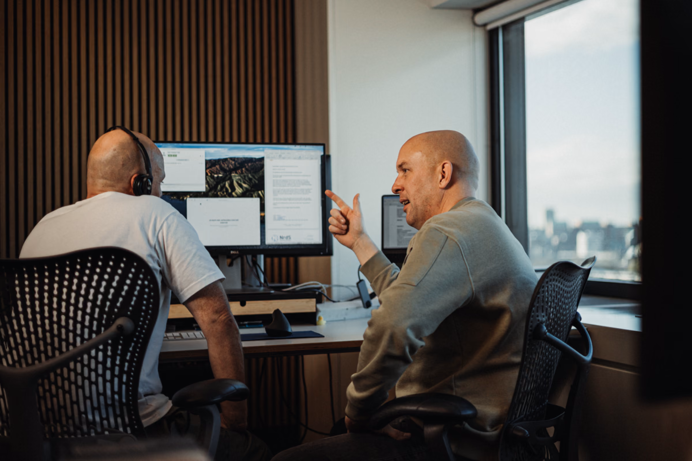 Two men talking at a desk in an office, one pointing at a computer screen with multiple windows open.