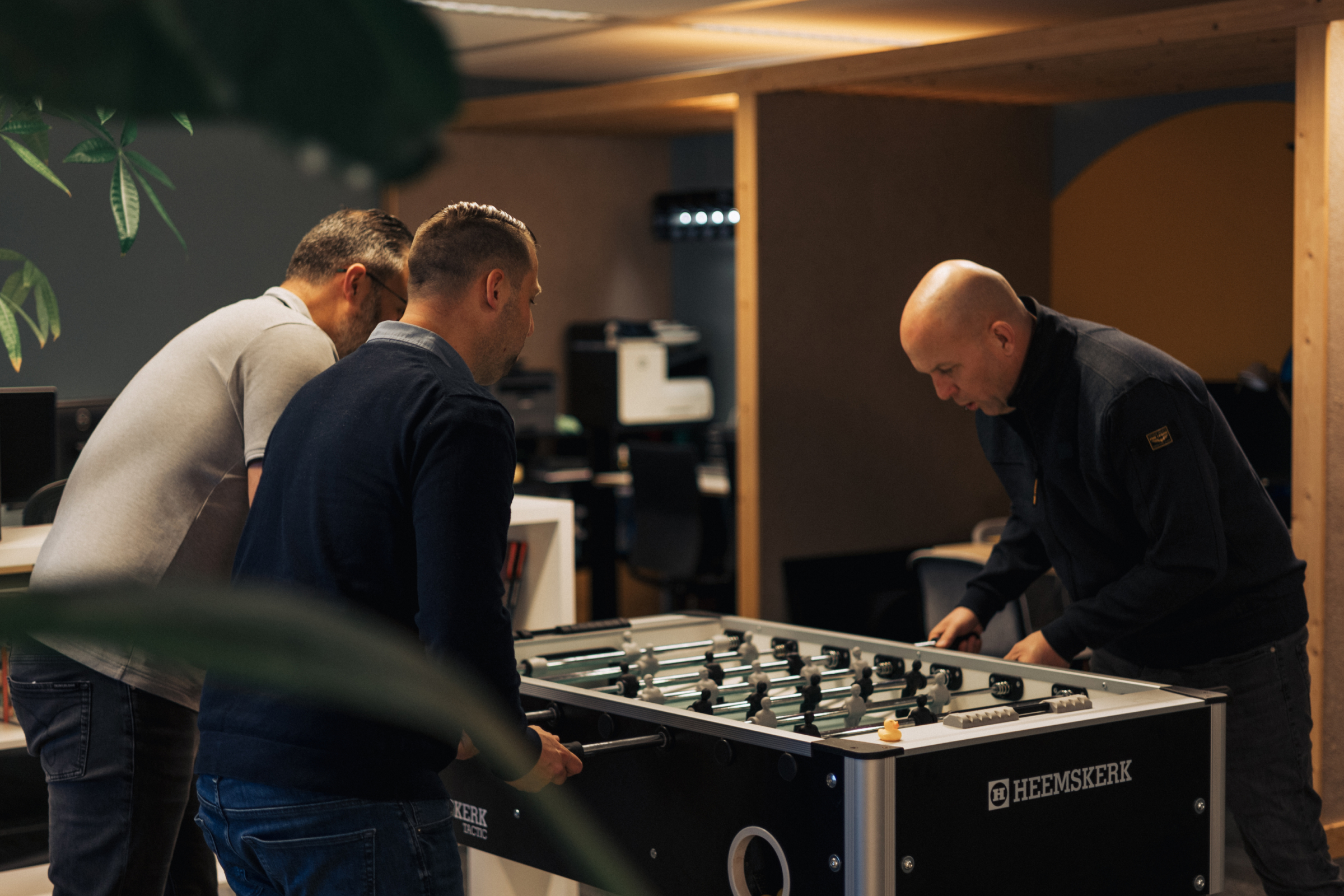 Three men playing foosball on a Heemskerk table in a cozy indoor space.