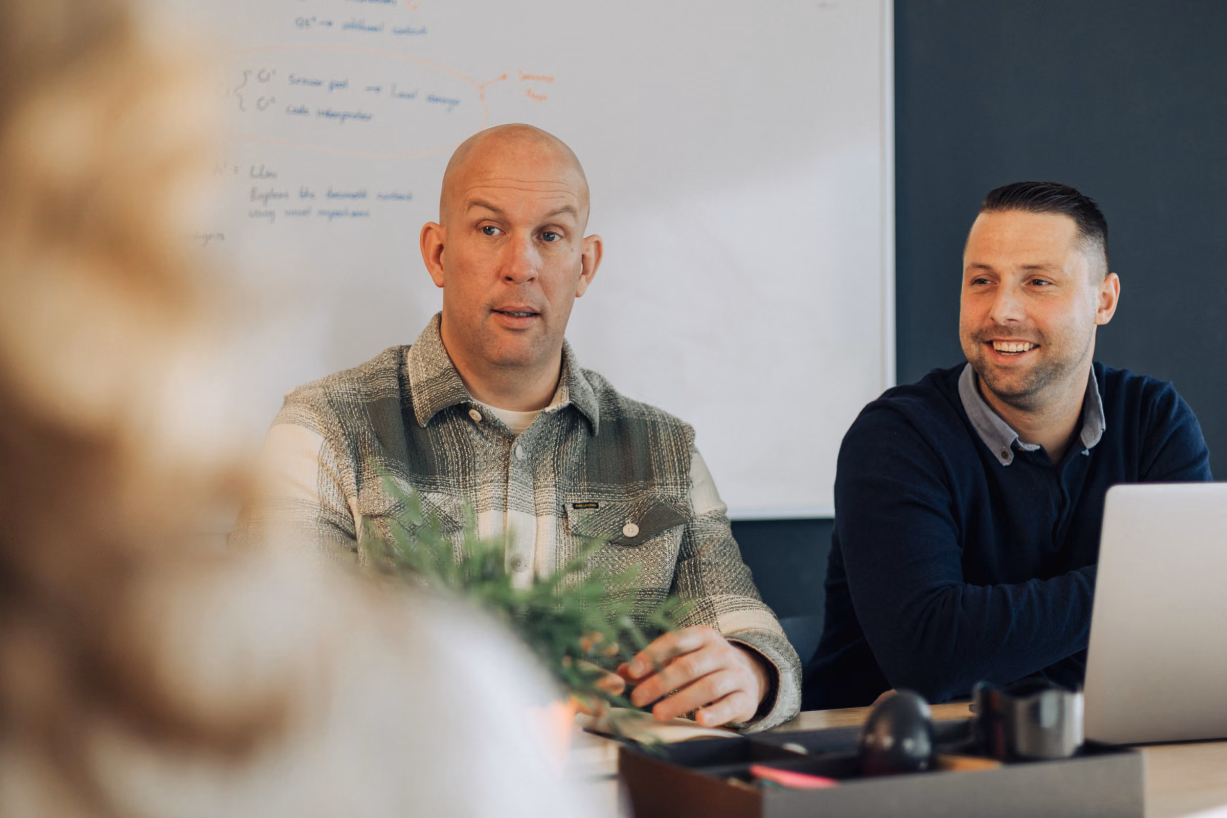 Two men sitting at a table in a meeting room with a whiteboard in the background.