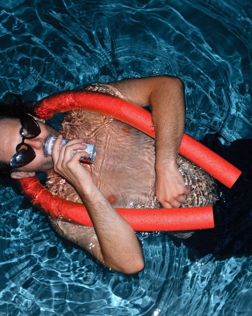 Someone floats in a pool on a red pool noodle, drinking from a Bowi can
