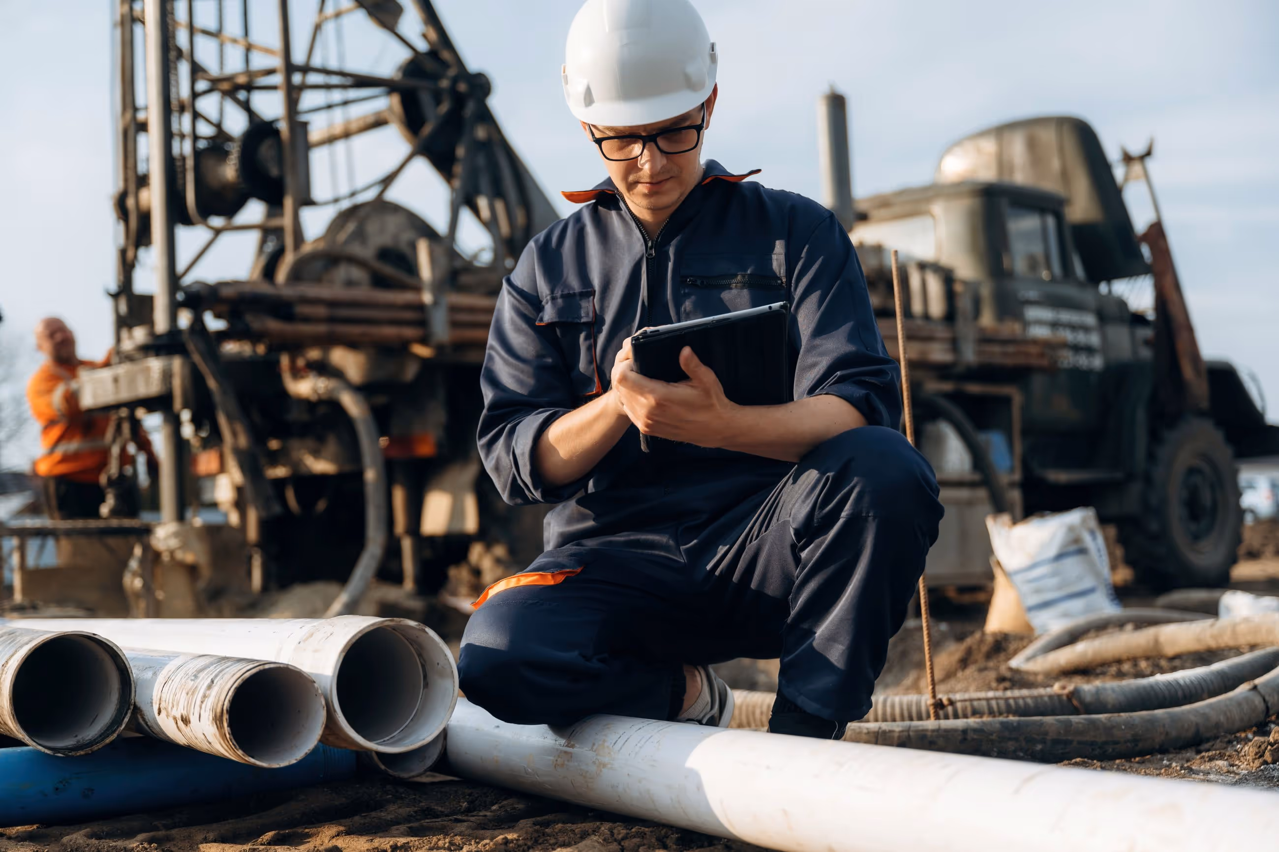 Construction worker kneels to inspect pipes, with machinery in the background