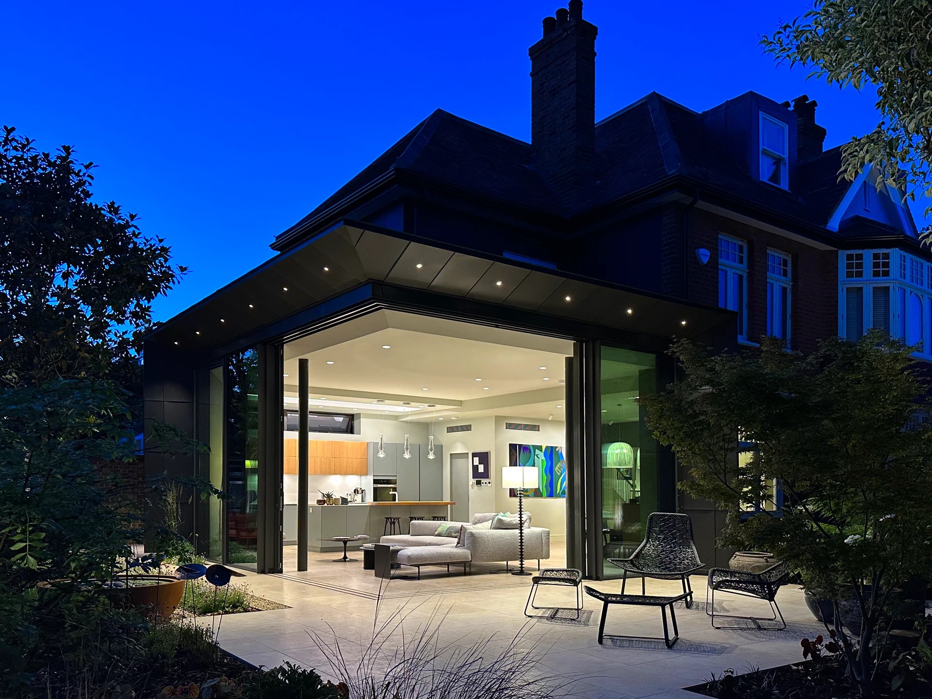 Contemporary glass extension with a black metal frame, seamlessly integrating with a traditional brick home and featuring an outdoor seating area.