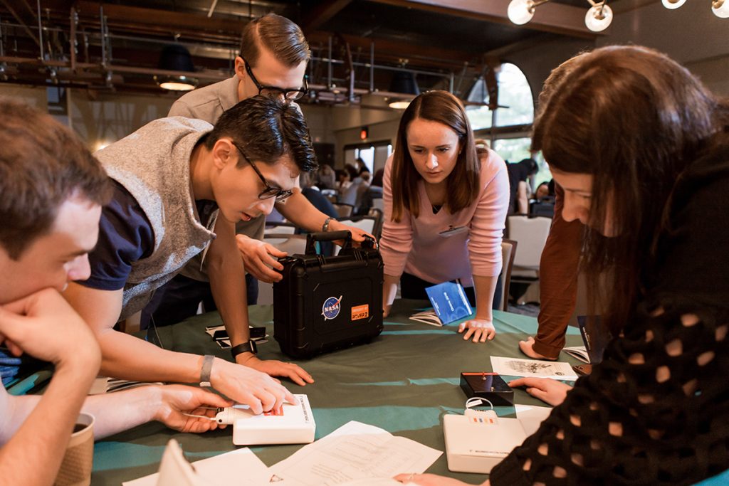 Vue d'ensemble d'un groupe de collaborateurs en entreprise autour d'une table avec des accessoires d'escape game déposés dessus : coffre en bois, cadenas, cartes, ambiance lumière naturelle dans une salle de réunion. Dynamisme et concentration mêlés.