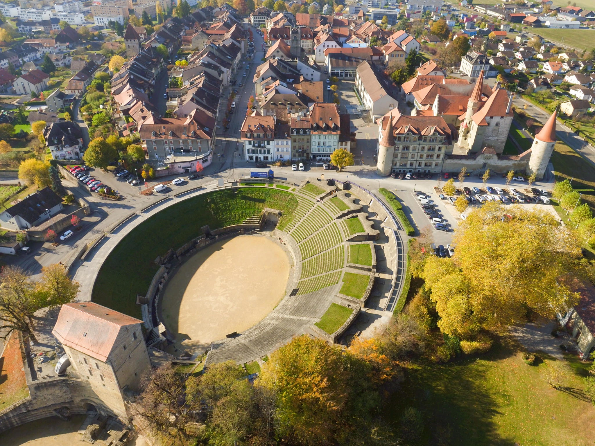 Vue des arènes romaines d'Avenches par une belle journée de printemps : gradins en pierre, ciel bleu, herbe verte au premier plan. Ambiance patrimoniale et sereine, lumière naturelle, pas de foule.