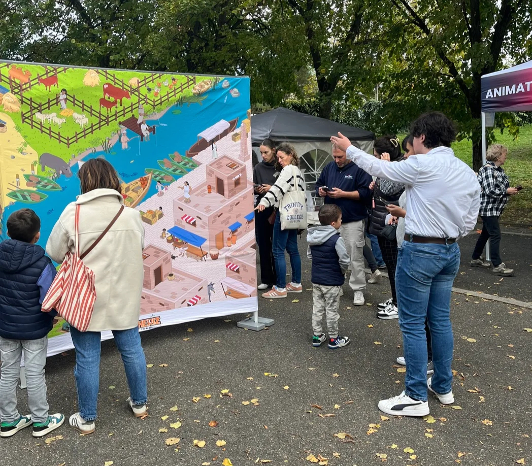 Vue d'un stand animé lors d'une fête de village vaudoise, groupe de participants de tous âges réunis autour d'un dispositif de jeu interactif en extérieur, badauds s'arrêtant pour regarder, ambiance festive et ensoleillée, décor de kermesse ou de festival.