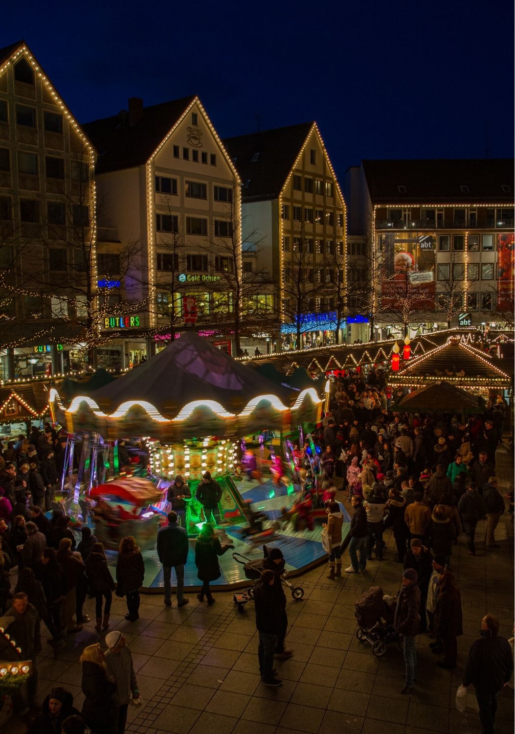 Vue d'ensemble d'une place de village vaudoise animée en soirée, lumières festives, stands ouverts, foule détendue, et dans le cadre un espace d'animation visible avec participants engagés autour d'un dispositif de jeu, ambiance chaleureuse de fête populaire estivale