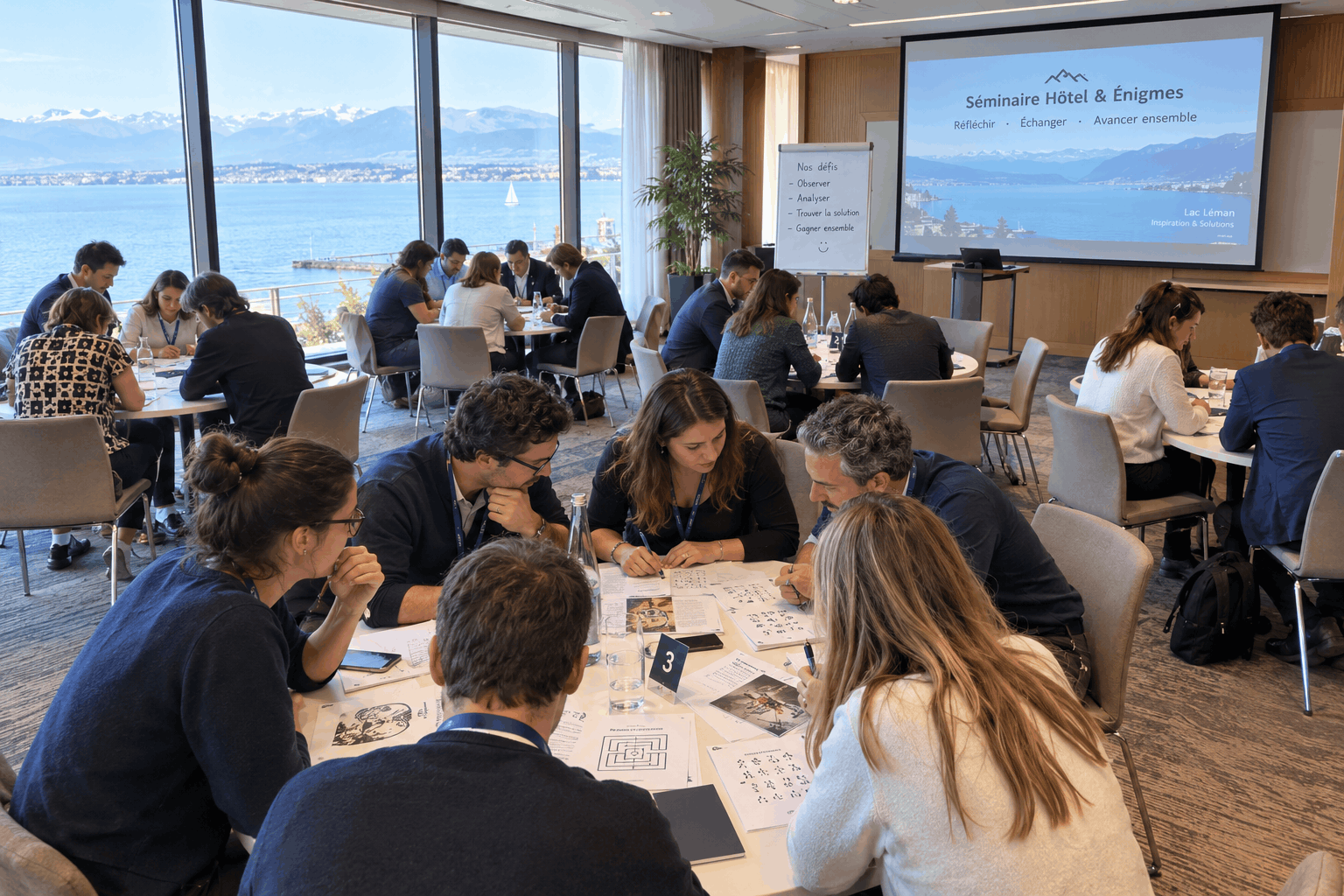 Salle de séminaire d'un hôtel avec vue sur le lac Léman : une trentaine de participants répartis en petites équipes autour de tables rondes, chacune penchée sur un coffre en bois et des documents d'énigmes. Ambiance studieuse et animée, lumière naturelle, paperboard et écran de plénière visibles en arrière-plan