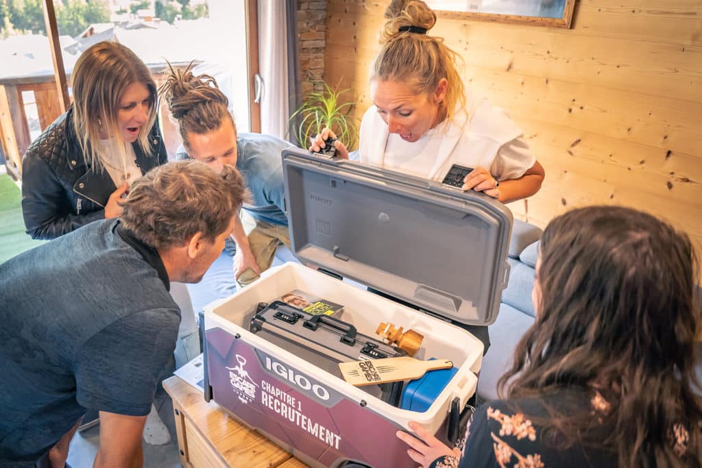 Groupe de cinq ou six amis dans un salon, autour d'une table basse, au moment où ils ouvrent une grande glacière : bouteilles de vin et de bière artisanale, saucisson, morceau de Gruyère et pain visibles. Visages réjouis, cadenas ouverts et feuilles d'énigmes éparpillés au premier plan, lumière chaude de soirée.
