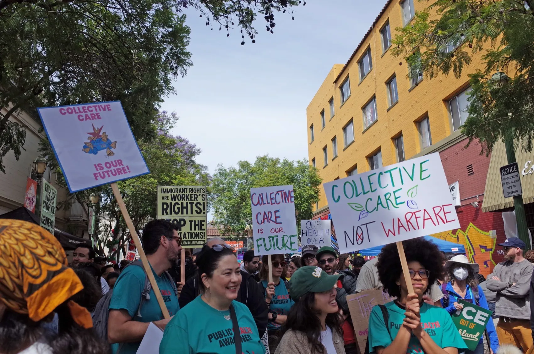 Protesters holding signs advocating collective care, immigrant rights, and peace in an urban setting.