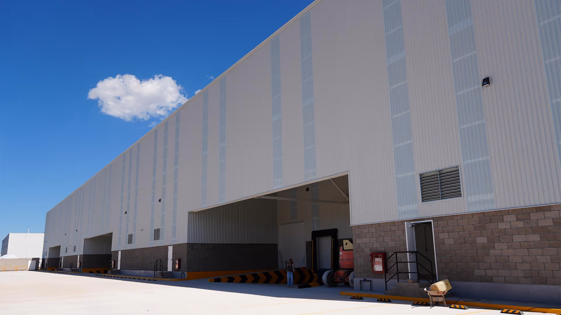 Vista exterior de un gran almacén industrial con muelles de carga y una persona de pie cerca de una de las bahías abiertas bajo un cielo azul claro.