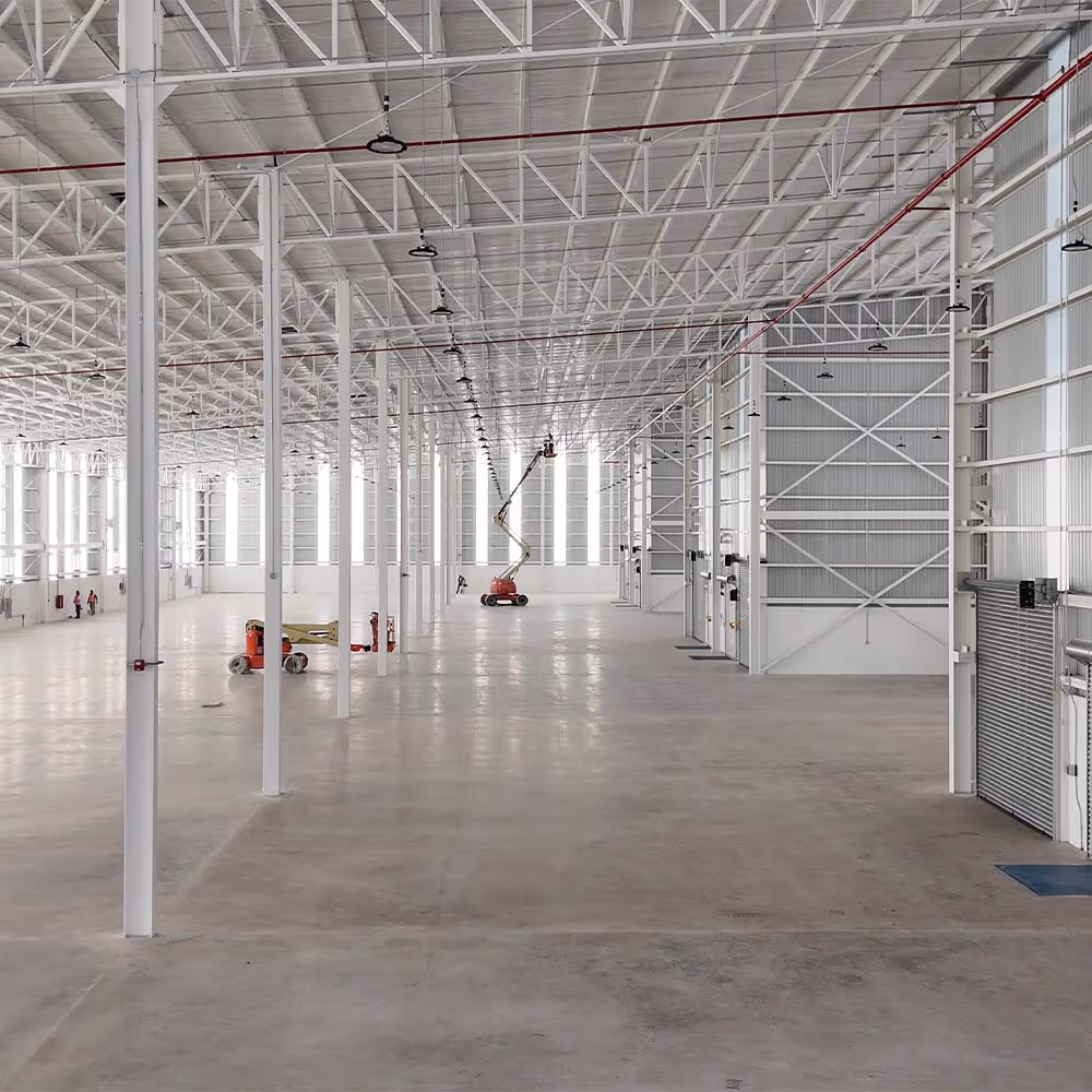Amplio almacén industrial vacío con vigas de acero blancas y un trabajador en una plataforma elevadora cerca del centro.