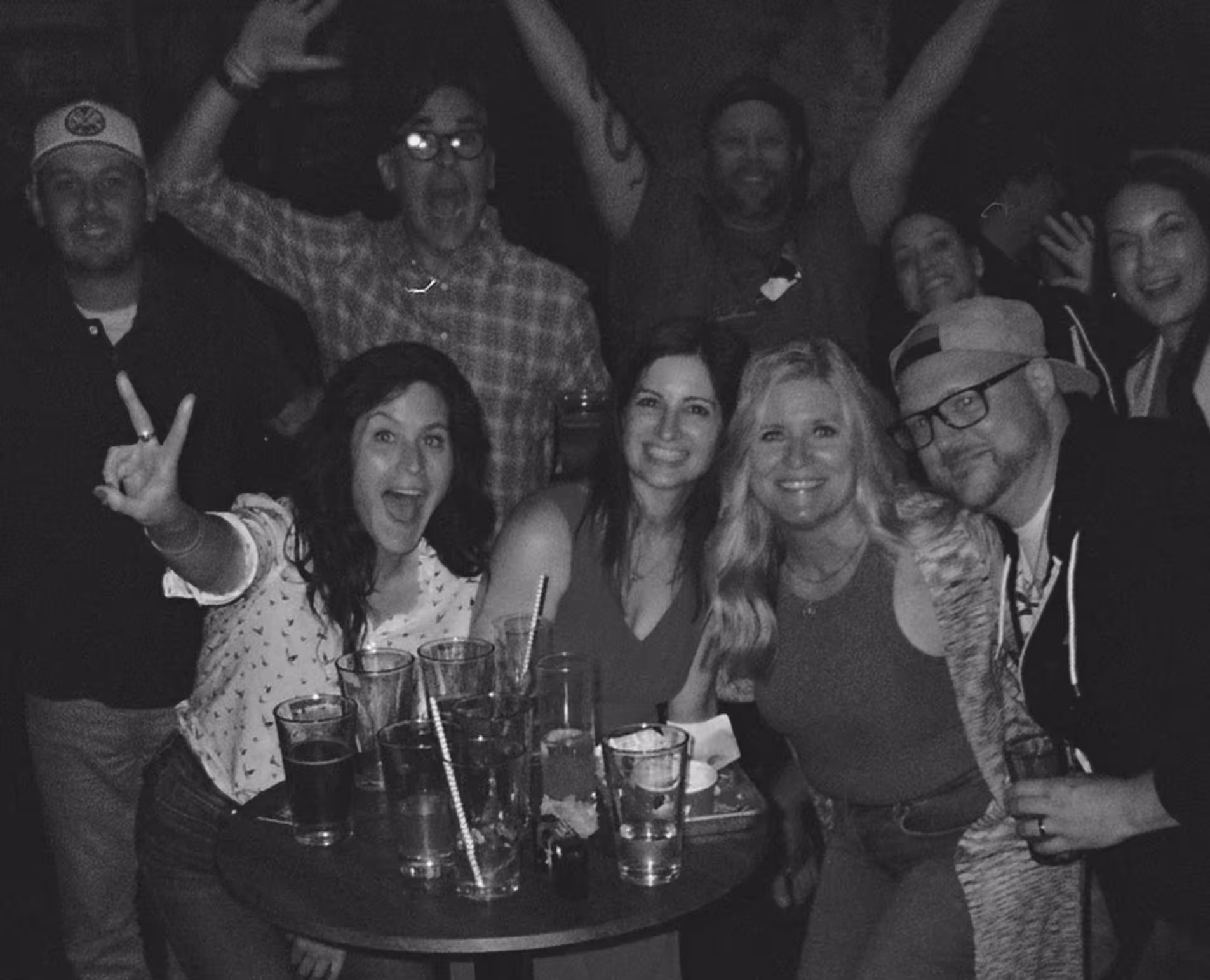 Group of smiling friends posing and making gestures around a small table with drinks in a nightlife setting.