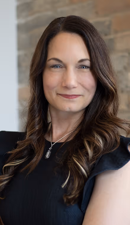 Woman with long wavy brown hair wearing a black top and a silver pendant necklace, smiling gently in front of a brick wall.