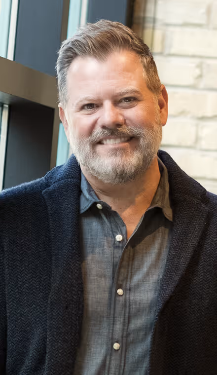 Smiling man with gray beard and short hair wearing a dark shirt and navy blue cardigan standing indoors near a window and brick wall.