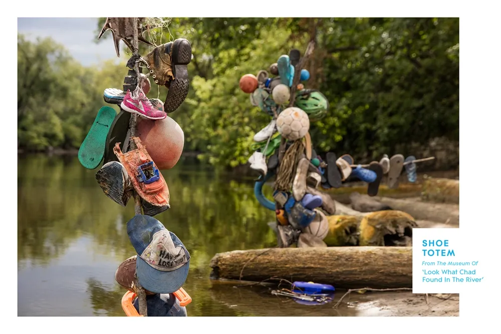 Two totem poles made from various shoes, sports balls, and other objects standing in a river surrounded by trees.