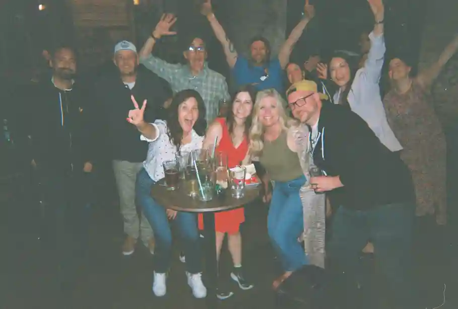 Group of friends gathered around a small table with drinks, smiling and posing for a photo in a dimly lit indoor setting.