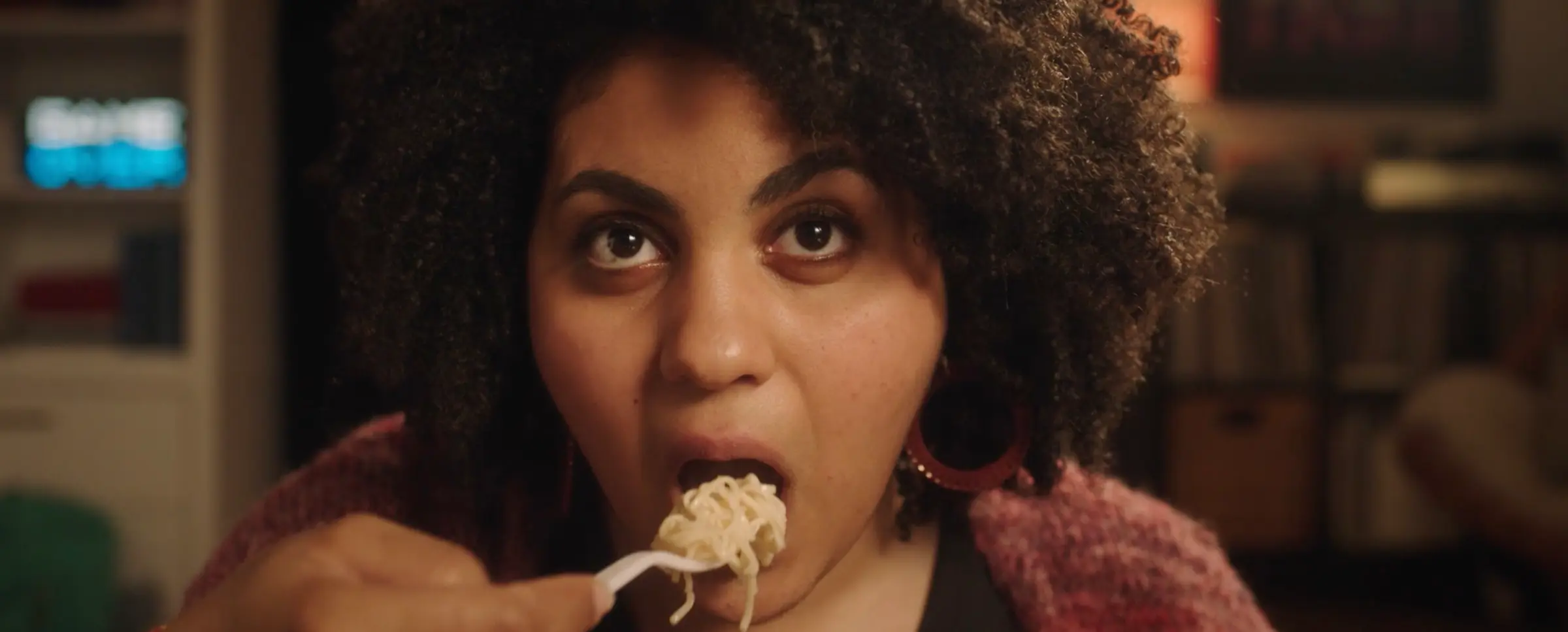 Close-up of a woman with curly hair eating noodles with a white fork indoors.