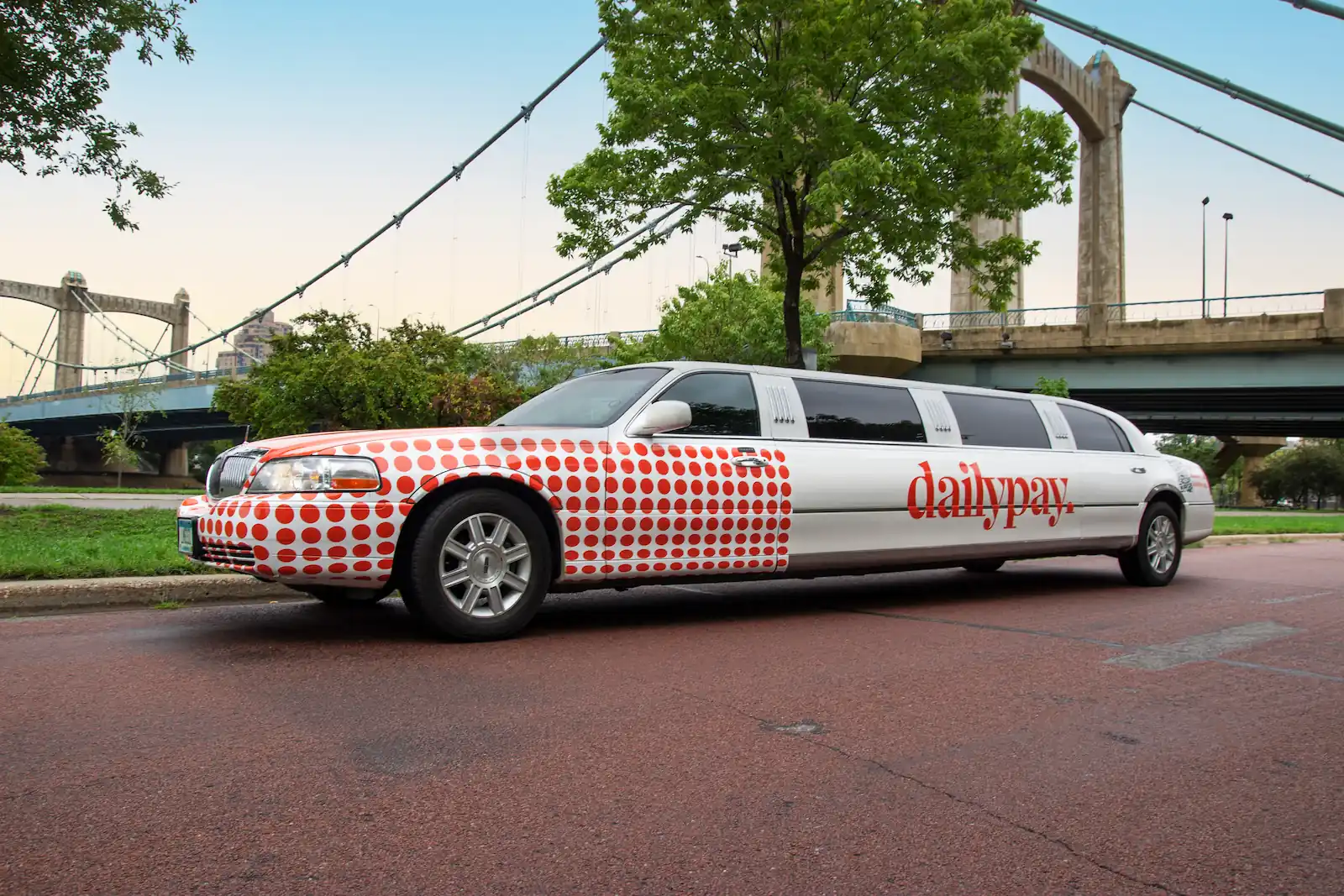 White stretch limousine with red polka dots and 'dailypay' logo parked on a street near a bridge and trees.