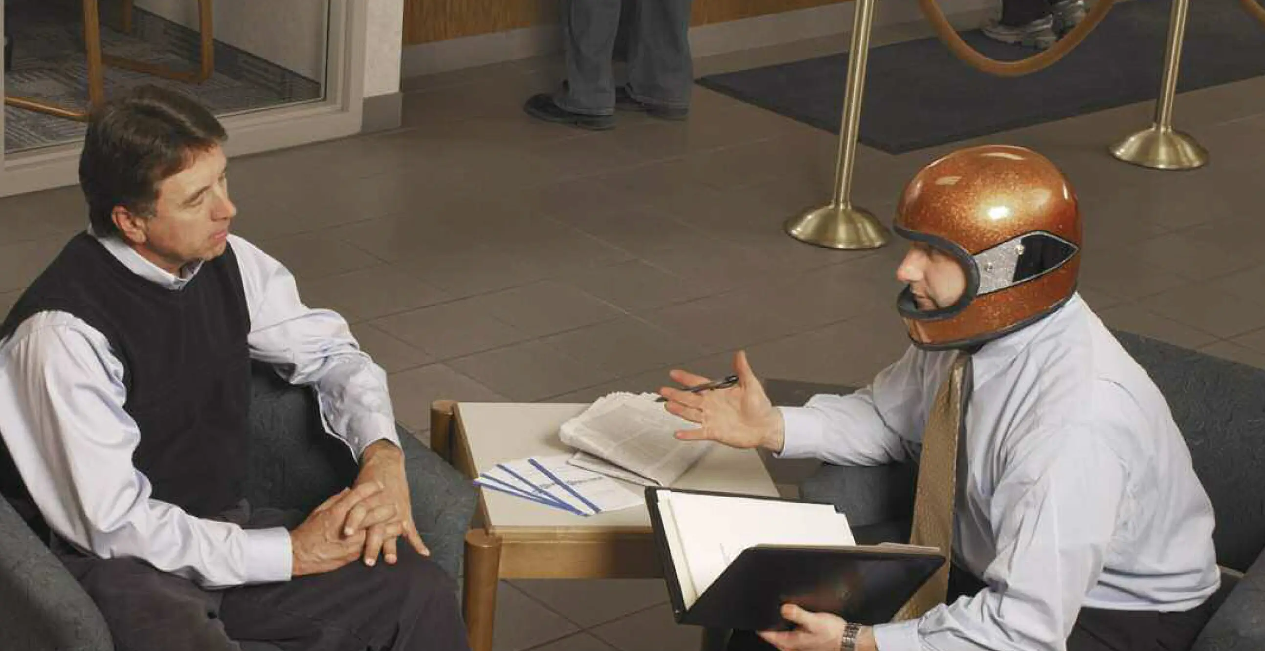 Two men sitting in an office talking, one wearing a shiny orange motorcycle helmet.