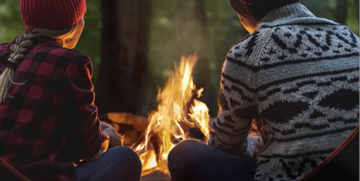 Two people sitting near a campfire in a forest, wearing warm sweaters and hats.