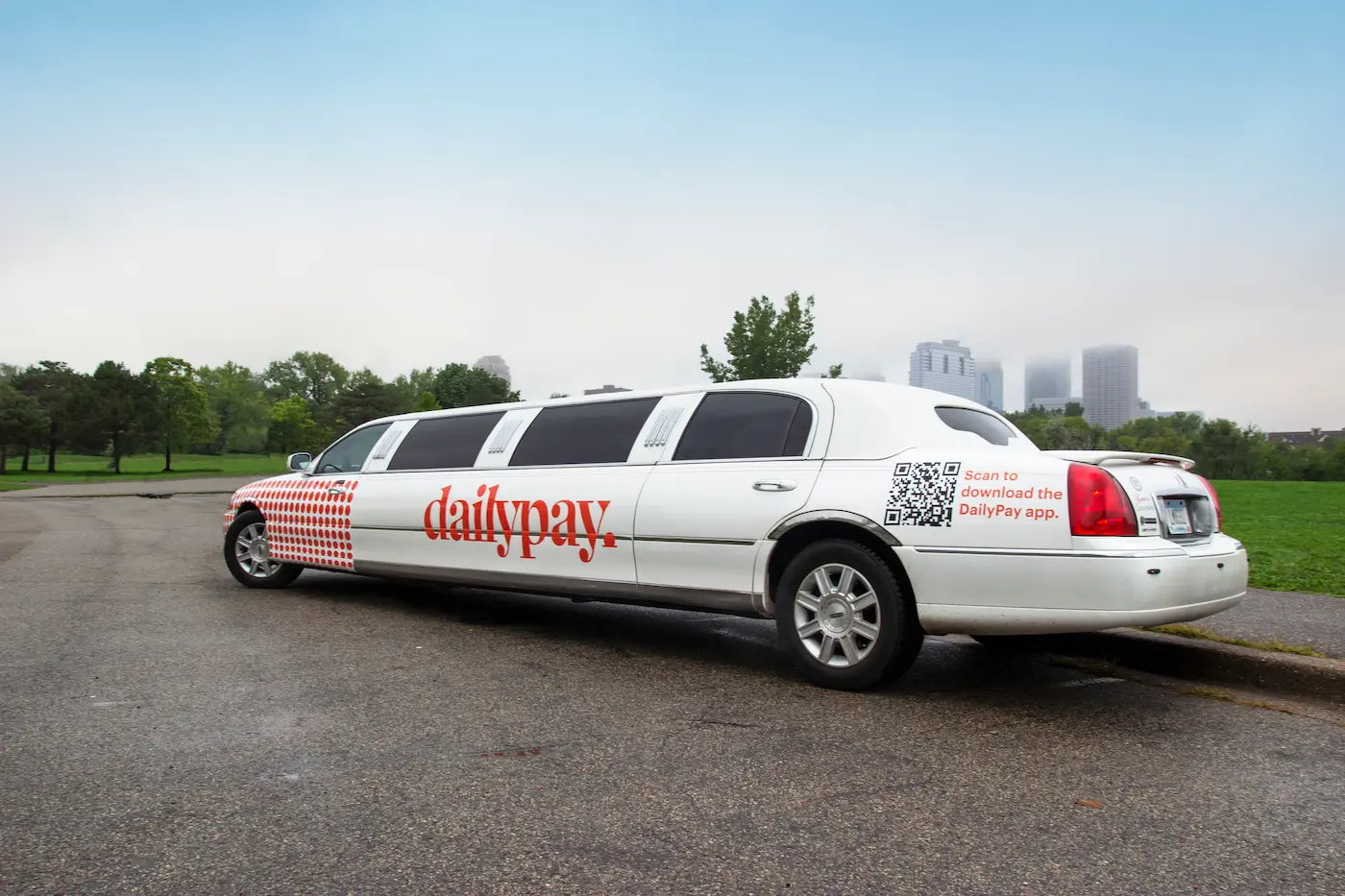 White stretch limousine with DailyPay branding and QR code parked on a street near a grassy park under a cloudy sky.