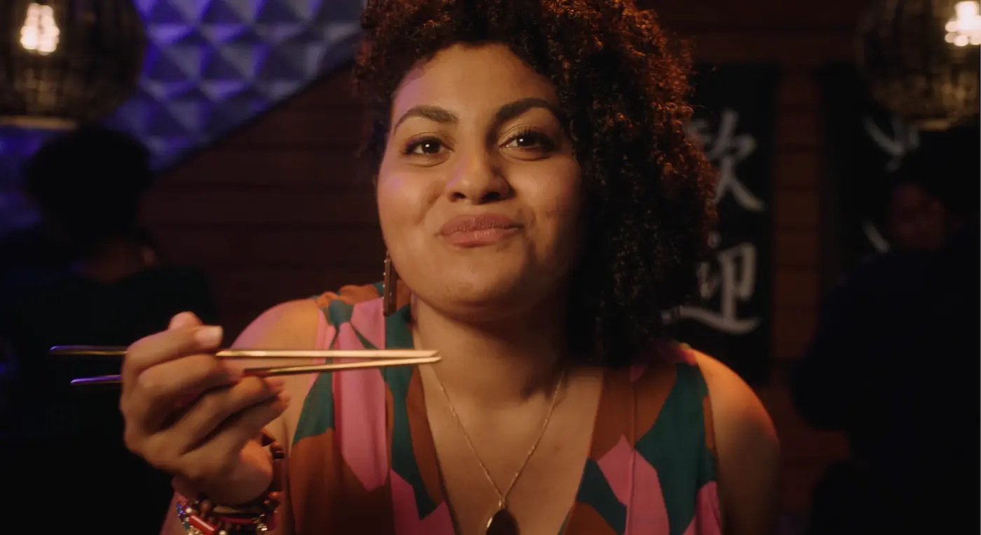 Woman with curly hair smiling and holding chopsticks in a dimly lit restaurant.