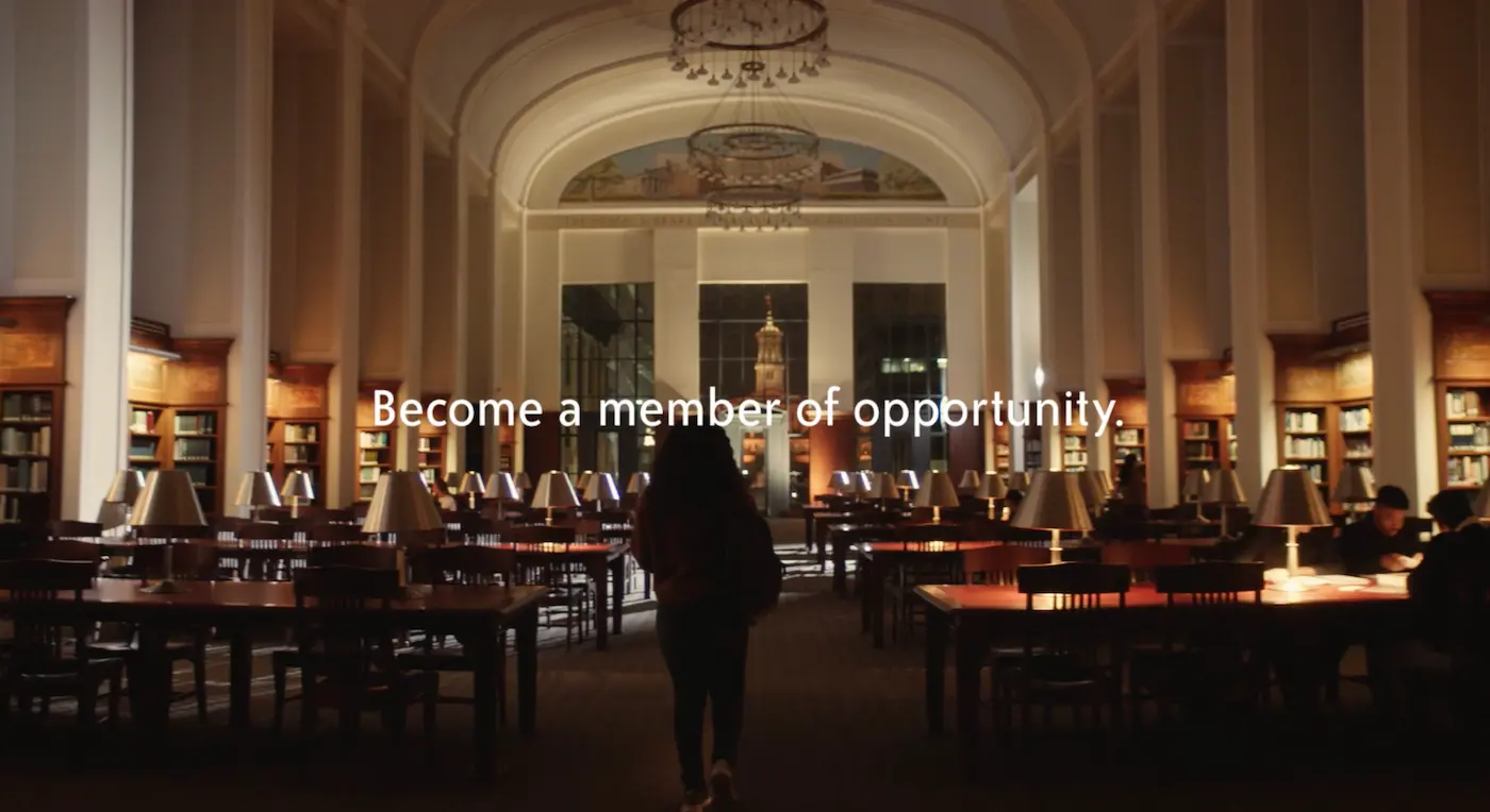 Person walking in a large, dimly lit library reading room with rows of tables and lamps, and text reading 'Become a member of opportunity.'