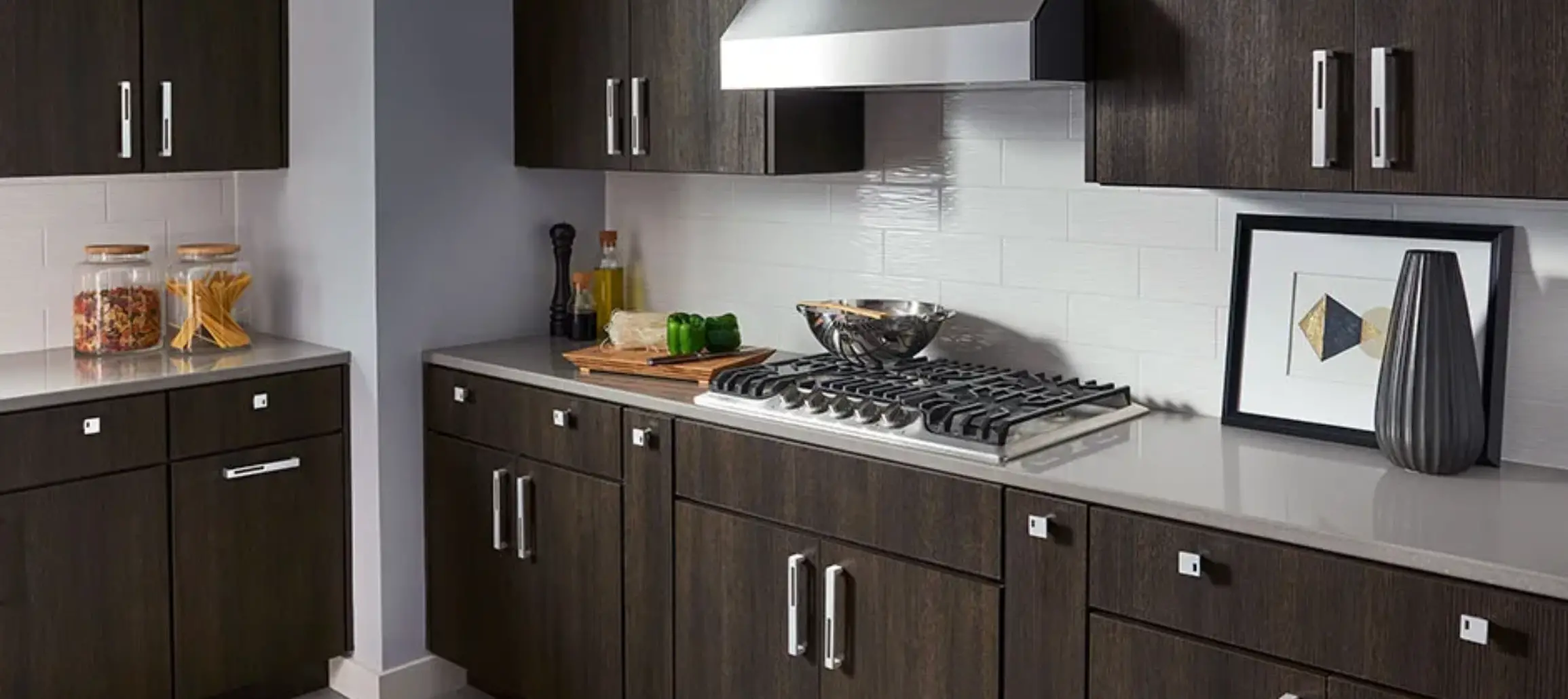Modern kitchen countertop with dark wood cabinets, a gas stove, cutting board with green bell peppers, glass jars with pasta, and decorative items.