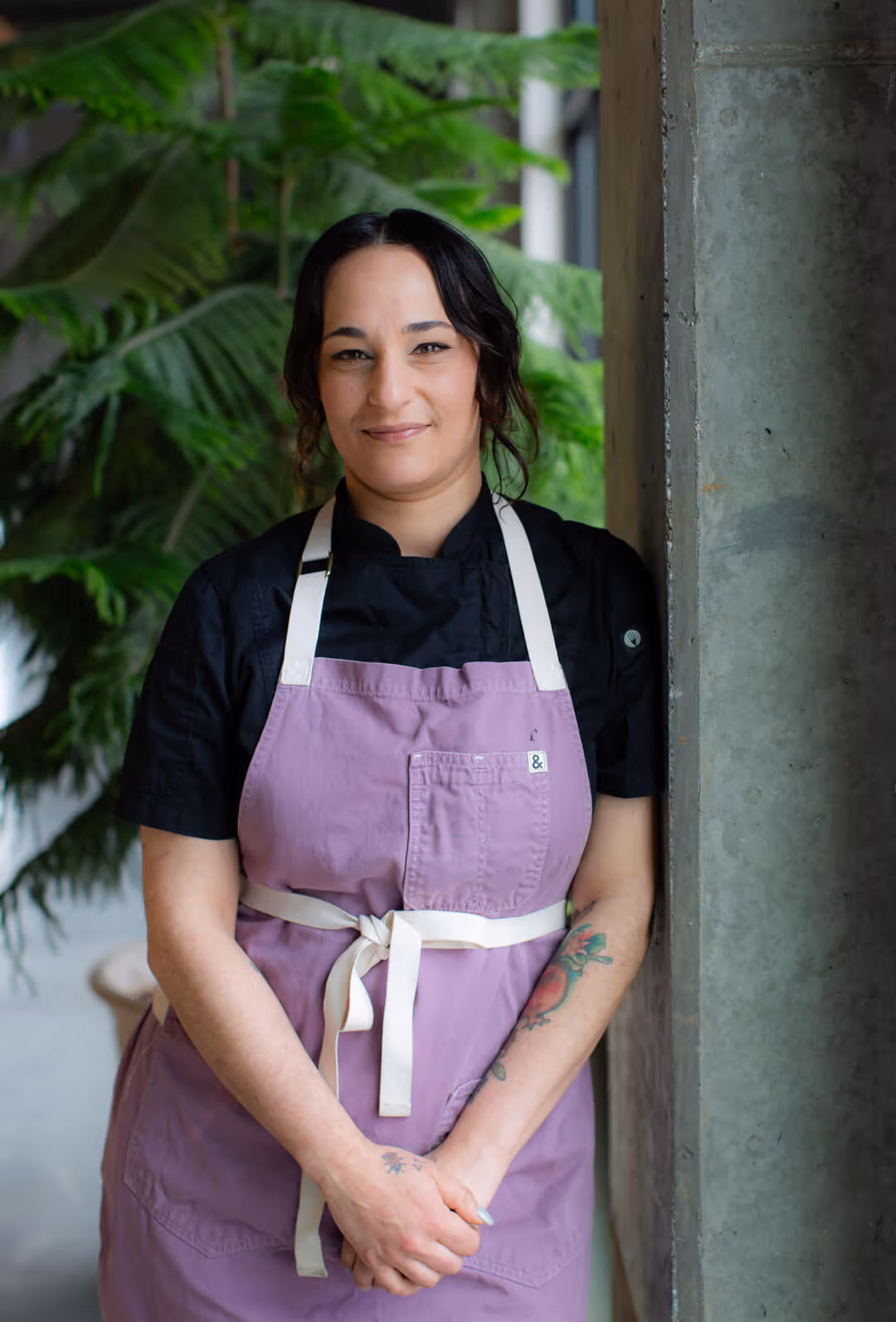 Danielle Grigorian wearing a black shirt and lavender apron smiling while leaning against a column