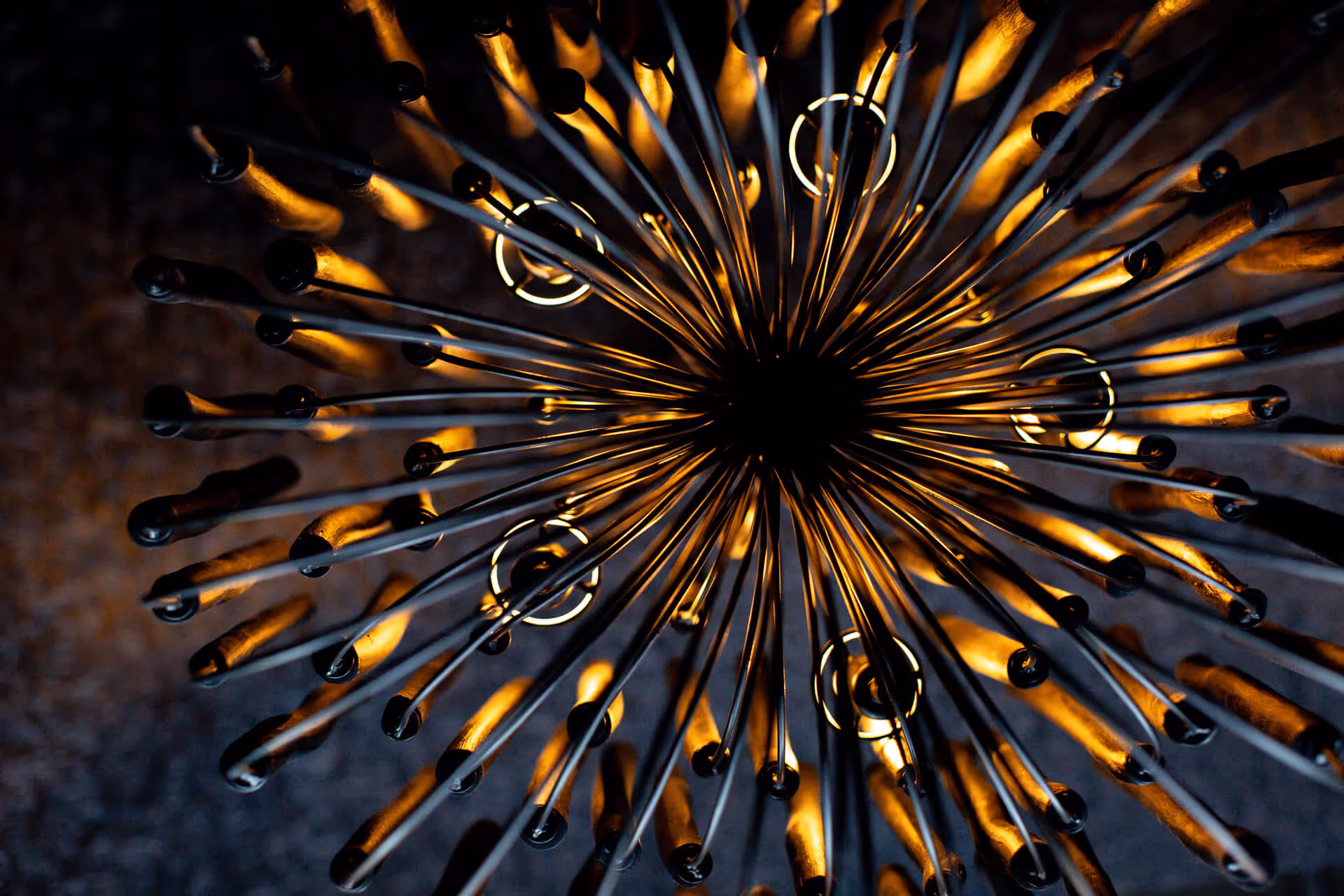 Abstract photo of the underside of a chandelier with dark metal and orange, glowing lights against a dark background