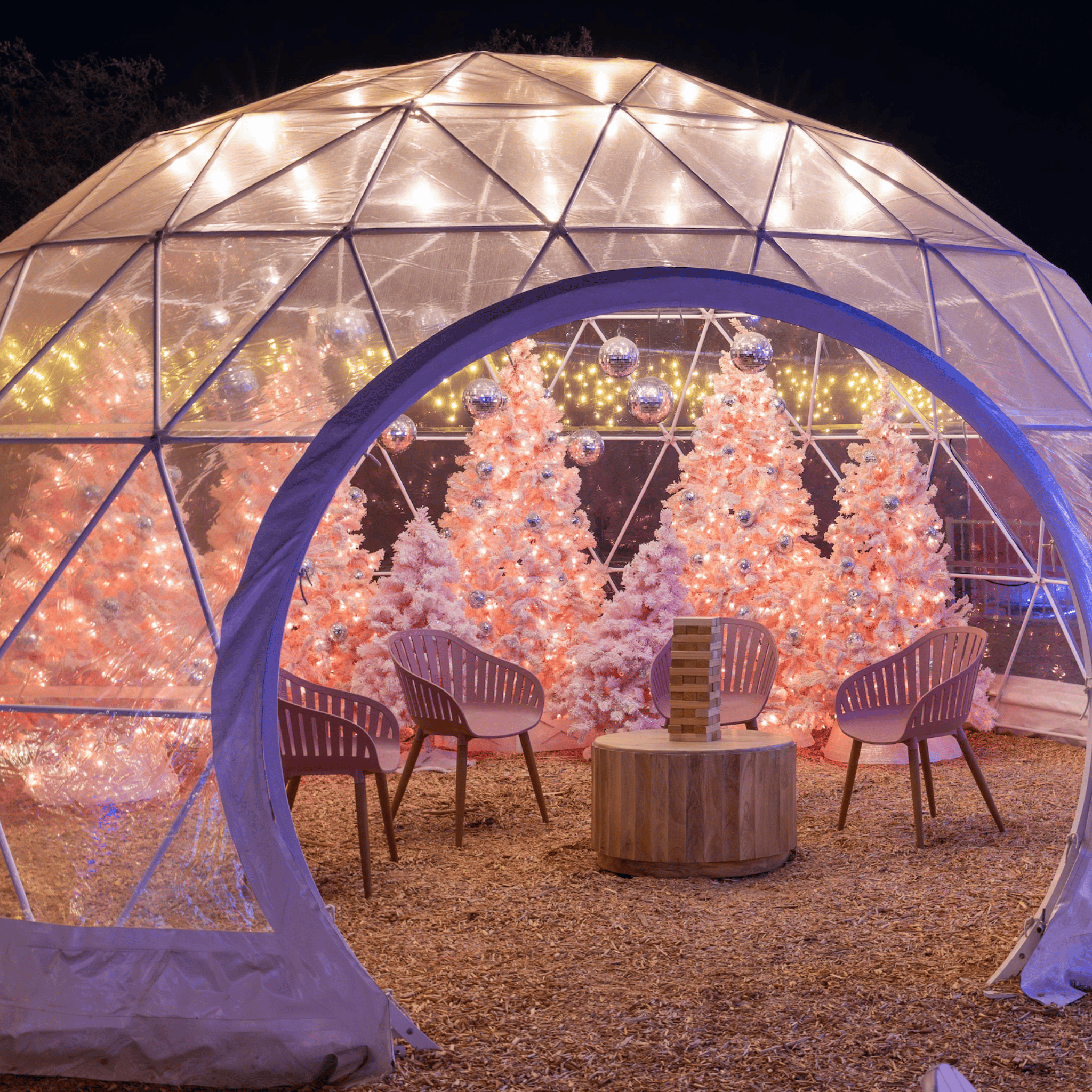 Lit transparent dome with pink Christmas trees, disco balls, four chairs, and wooden table with Jenga game.