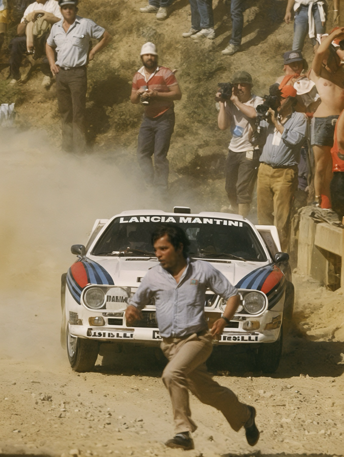 Une Lancia 037 Martini déboule sur une piste de terre, soulevant un nuage de poussière, tandis qu’un spectateur court pour éviter la trajectoire pendant un rallye du Groupe B en 1984.