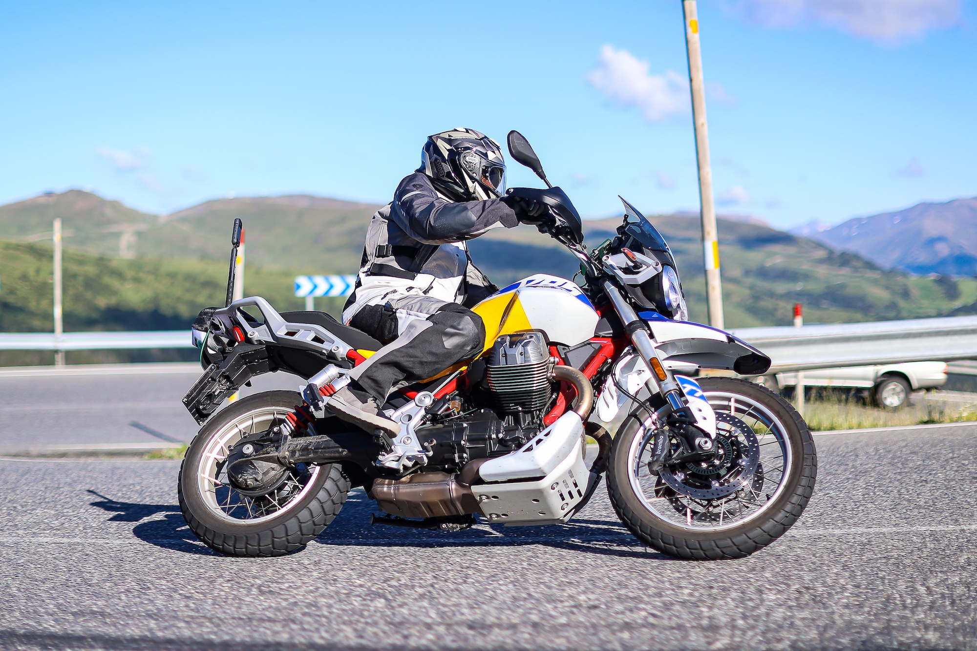 Moto Guzzi V85 TT jaune et bleue avec son pilote à un arrêt en montagne, une autre moto en arrière-plan.