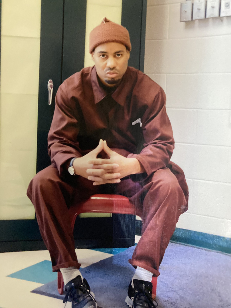 Yem, sitting on a chair with his hands folded, wearing a hat and jumpsuit.