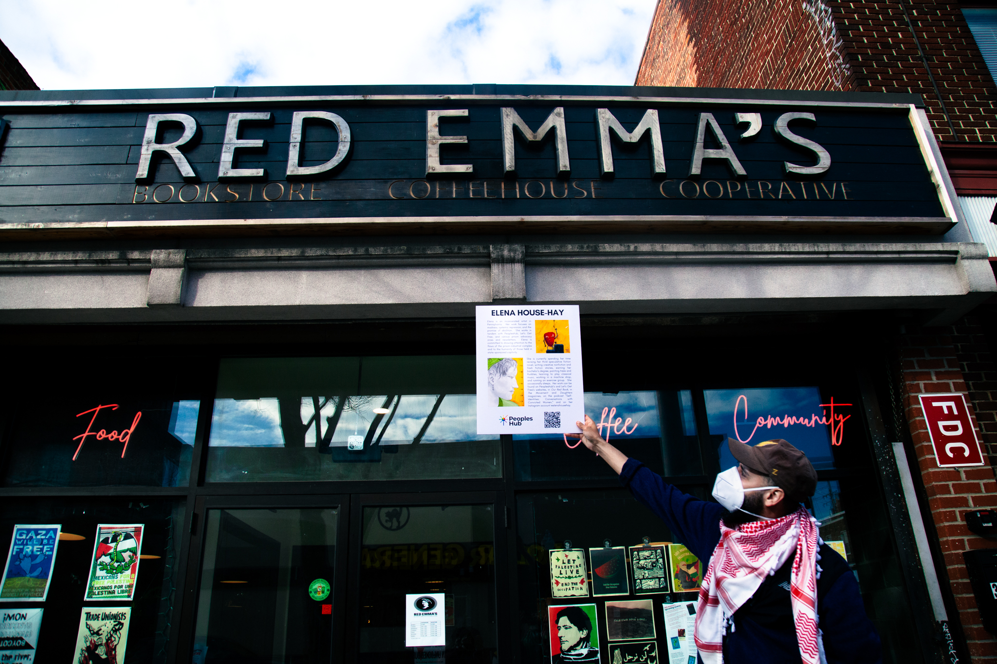 A person in a mask and hat holds up the exhibit sheet for Elena House-Hay outside of Red Emma's bookstore, coffee house and cooperative