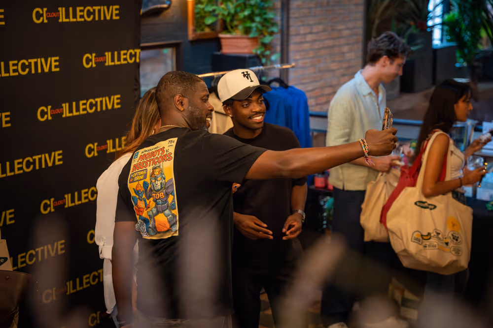 Group of people socializing indoors with one man taking a selfie in front of a black backdrop with yellow text.