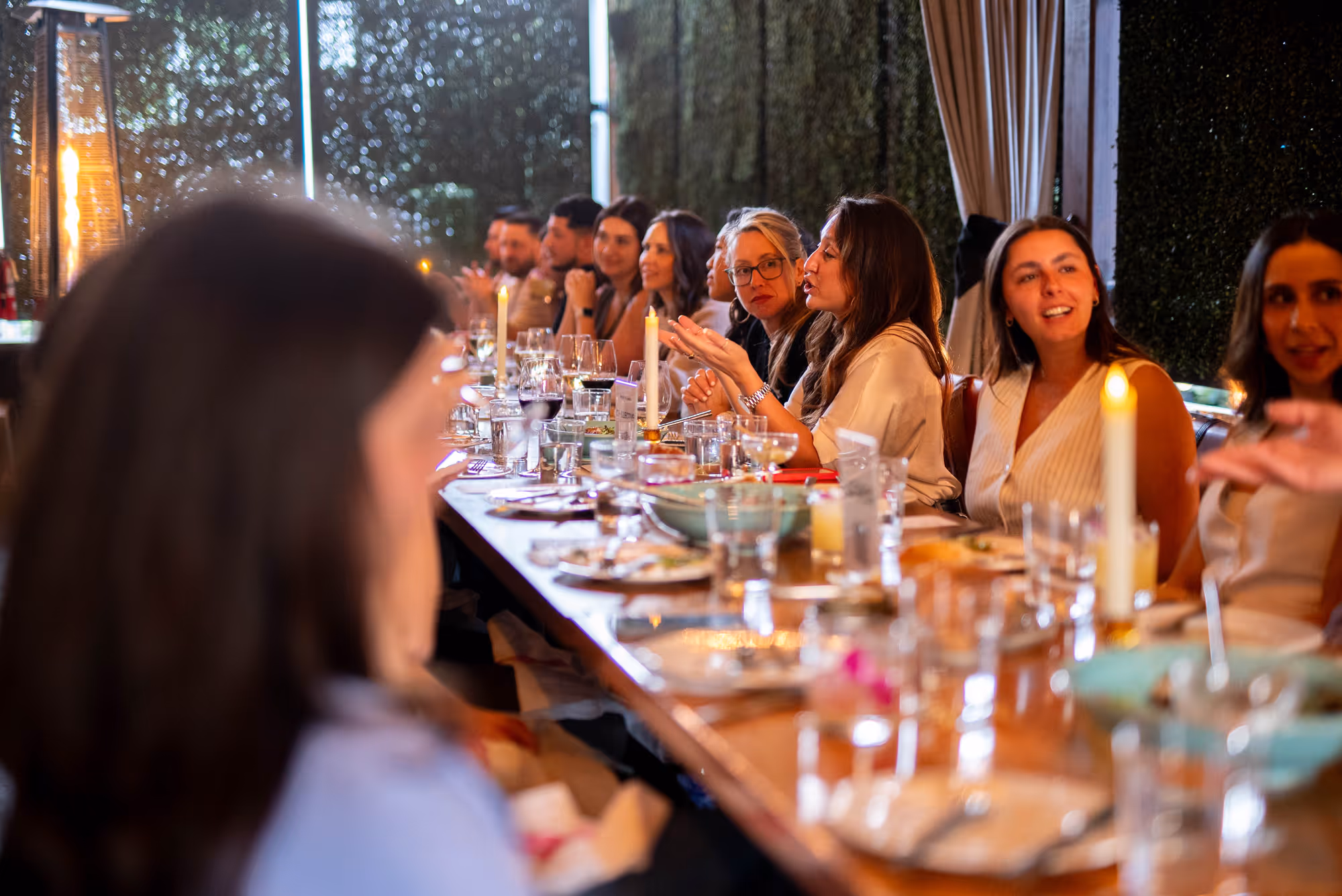 Group of people seated at a long table having a lively dinner conversation with candles lit.