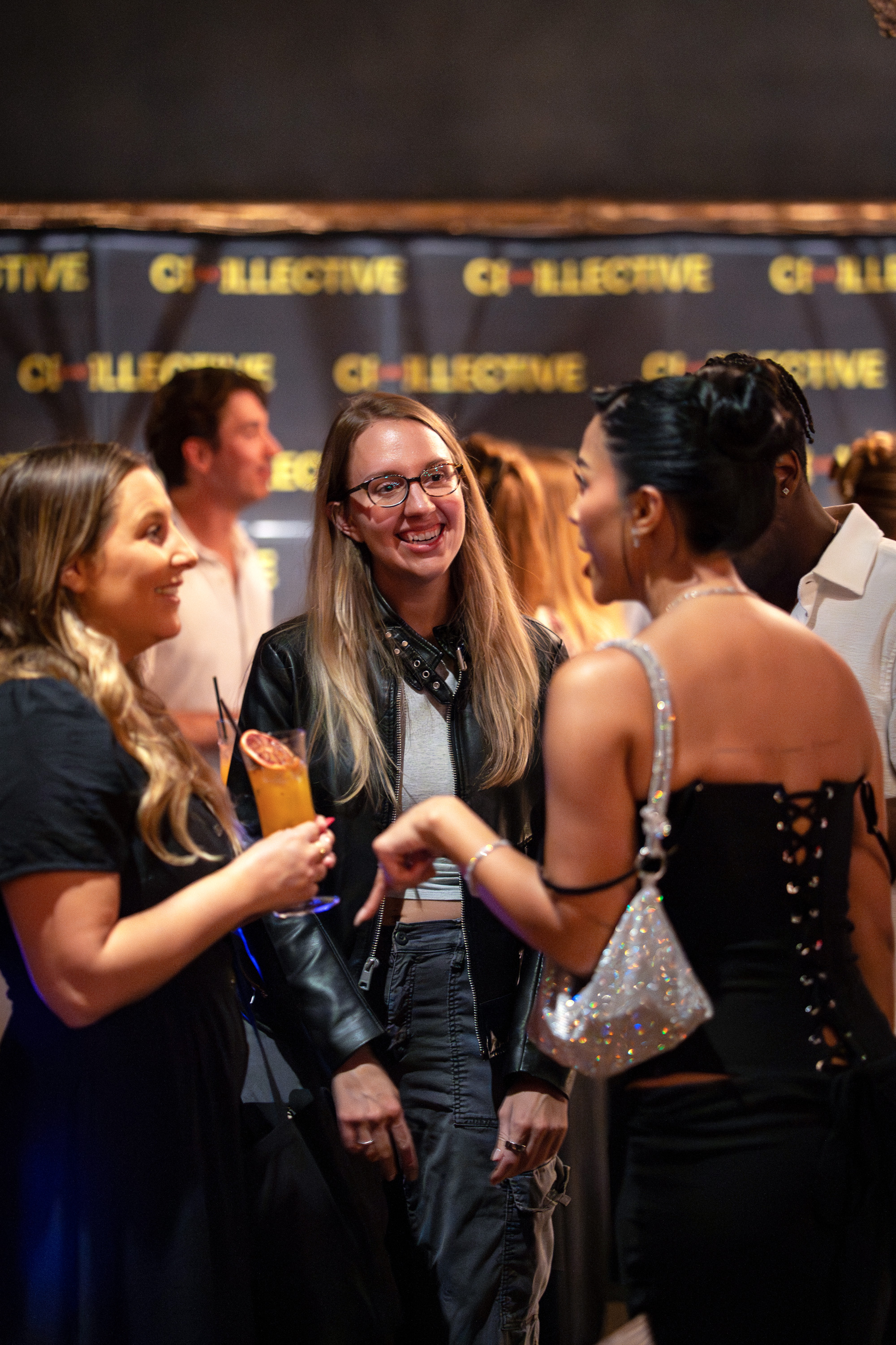Three women chatting at a social event with one holding a cocktail and smiling.