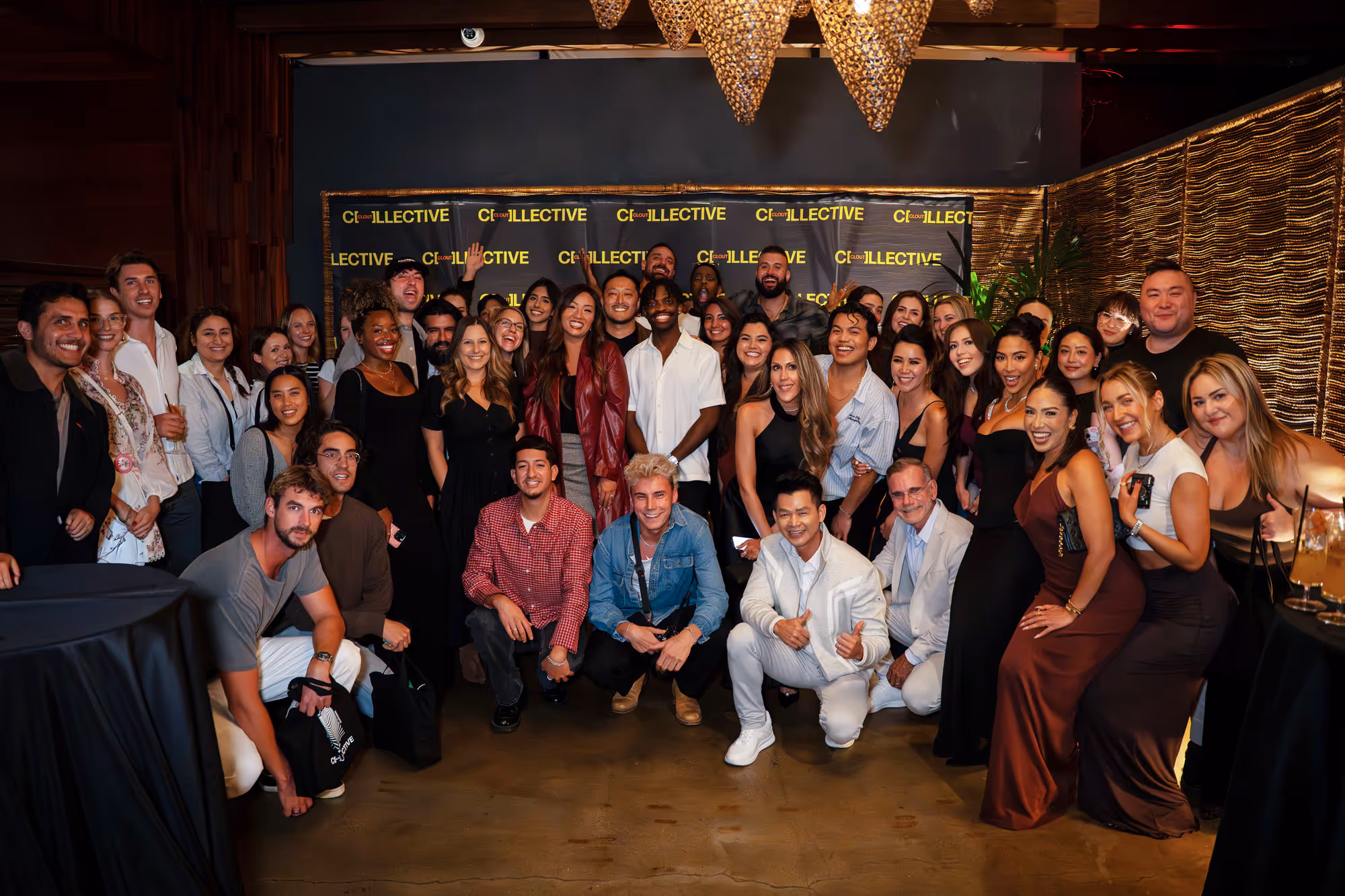 Large group of diverse people smiling and posing together in front of a Clout Collective backdrop at an indoor event.