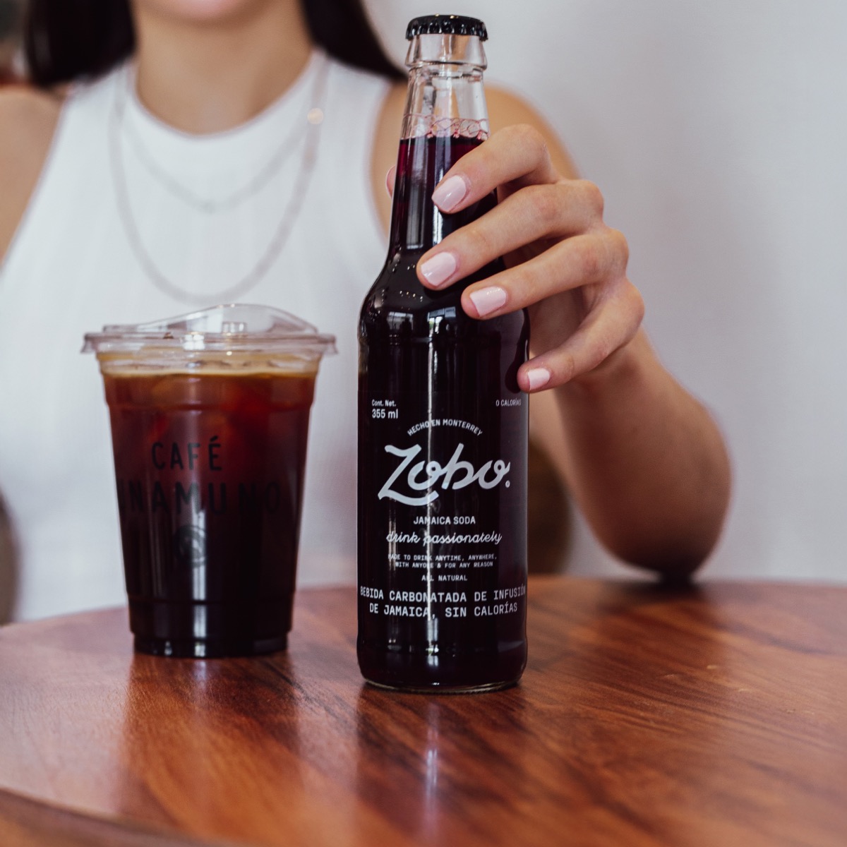 alguien agarrando una botella de vidrio de 355ml ZOBO en una mesa de madera junto a un café de CAFE UNAMUNO