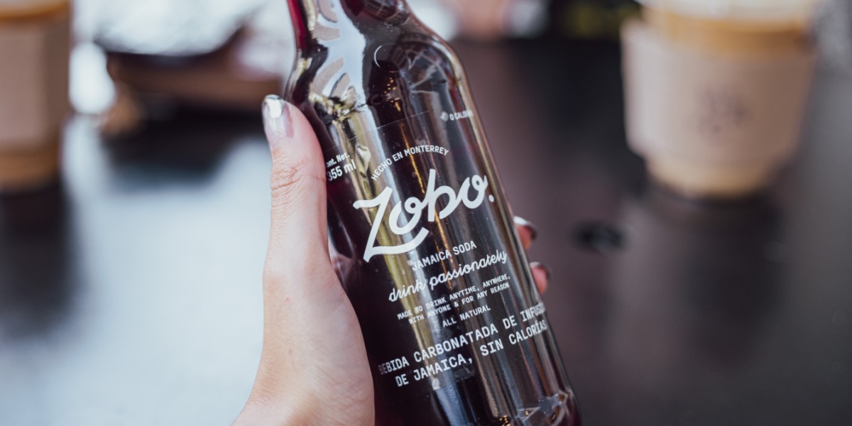 Hand holding a dark glass bottle of Zobo Jamaica soda with a blurred background.