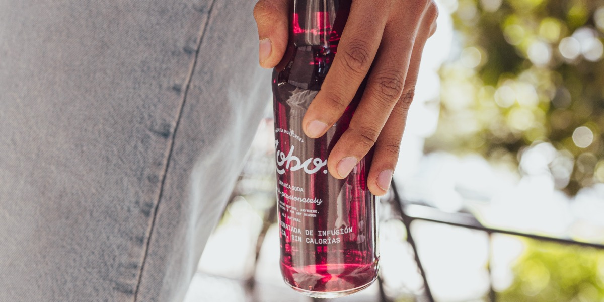 Close-up of a hand holding a glass bottle of Zobo Jamaica soda with blurred outdoor background.