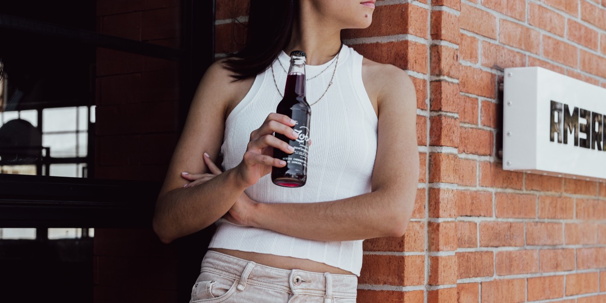 Person in a white sleeveless top holding a dark glass bottle, standing against a brick wall.