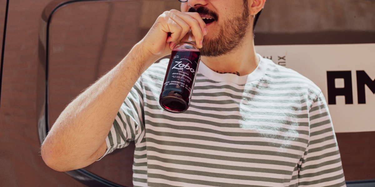 Man with glasses and beard drinking from a bottle of Zobo Jamaica soda, wearing a white and grey striped shirt.