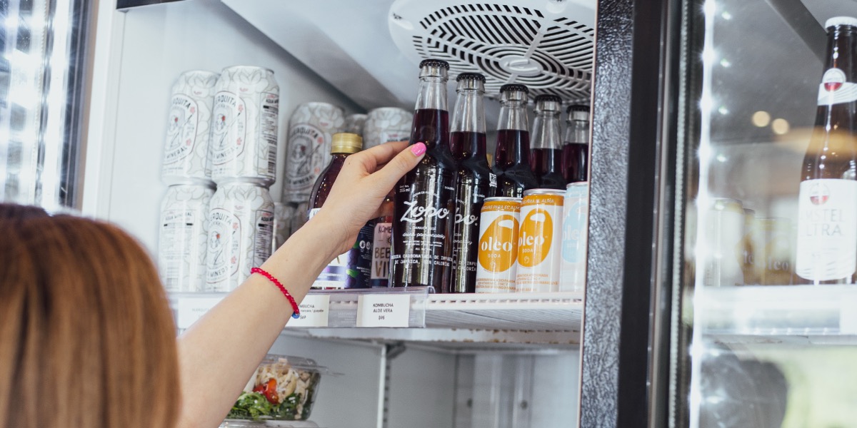 Person's hand with pink nail polish reaching for a dark glass bottle on a fridge shelf with assorted beverages including canned drinks and bottled kombucha.
