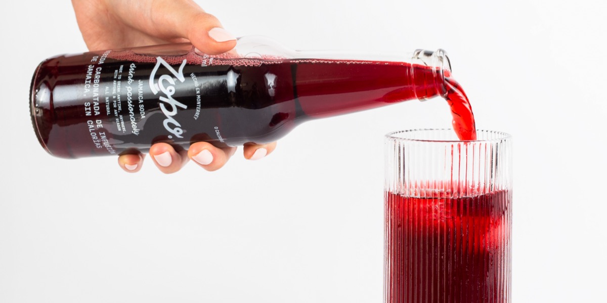 Hand pouring red soda from a Zesto glass bottle into a ribbed glass filled with ice.