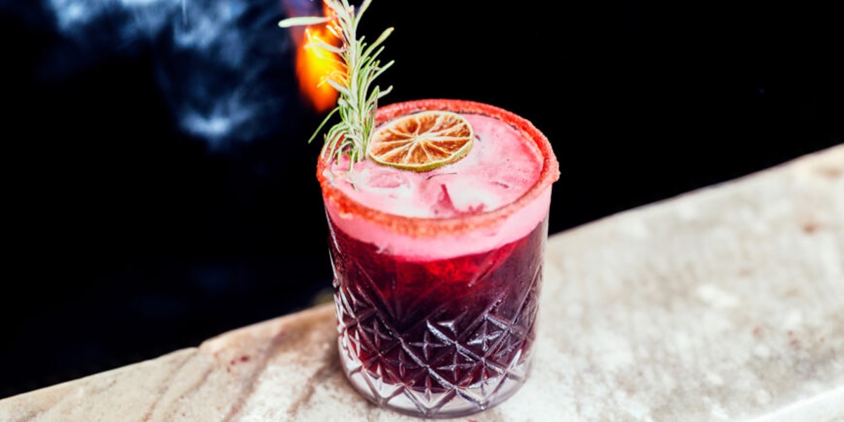Red cocktail in a textured glass with a salted rim, garnished with a dried lime slice and a sprig of rosemary on a stone surface.