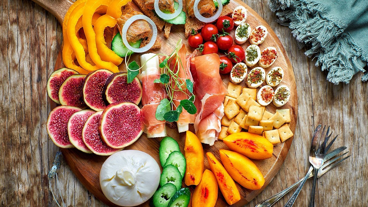 Wooden platter with sliced figs, yellow bell pepper rings, cucumber slices, cherry tomatoes, deviled eggs, crackers, prosciutto-wrapped cheese, burrata cheese, and thin sliced peaches on a rustic wooden table.