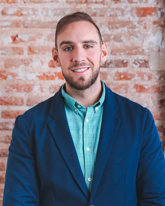 Smiling professional in navy blazer and teal shirt against brick wall